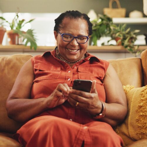 A black woman sits on a couch with a smile and looks at her phone.